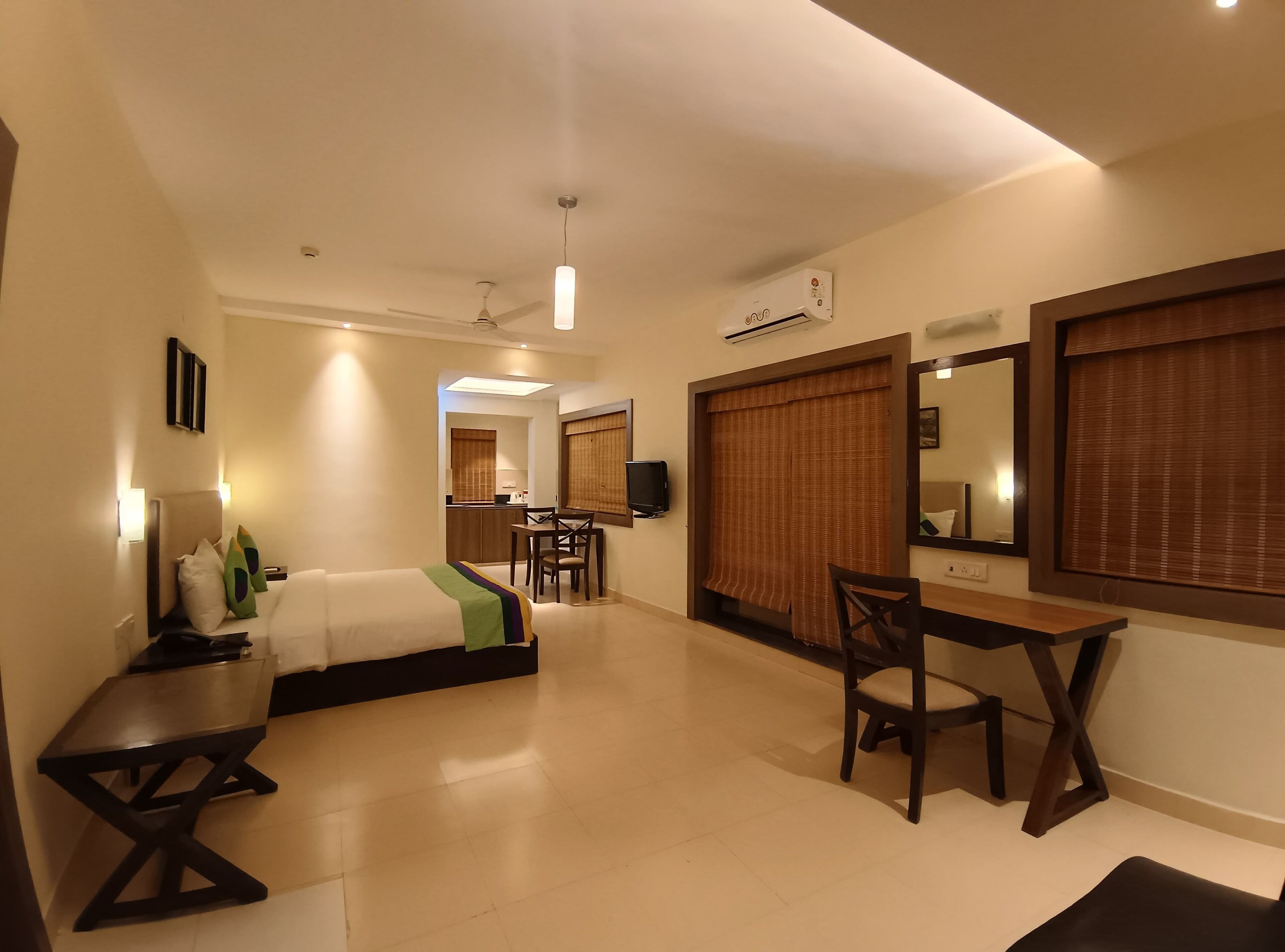 A hotel room with a bed, a desk, a chair, a mirror, and a kitchenette.