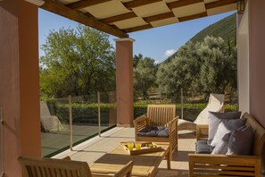 Un espacio de estar al aire libre techado con mobiliario de madera y una barrera de vidrio, con vistas a un paisaje montañoso con árboles.