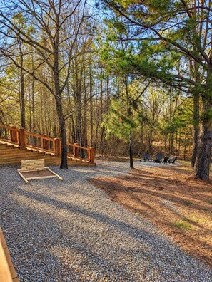 A forested area with a wooden deck, a small seating area with chairs, and a gravel ground. - Lofted Pines, Hot Tub, Foosball, Air Hockey, Corn Hole, Horse Shoes, Table Arcade, Tetherball (Broken Bow)