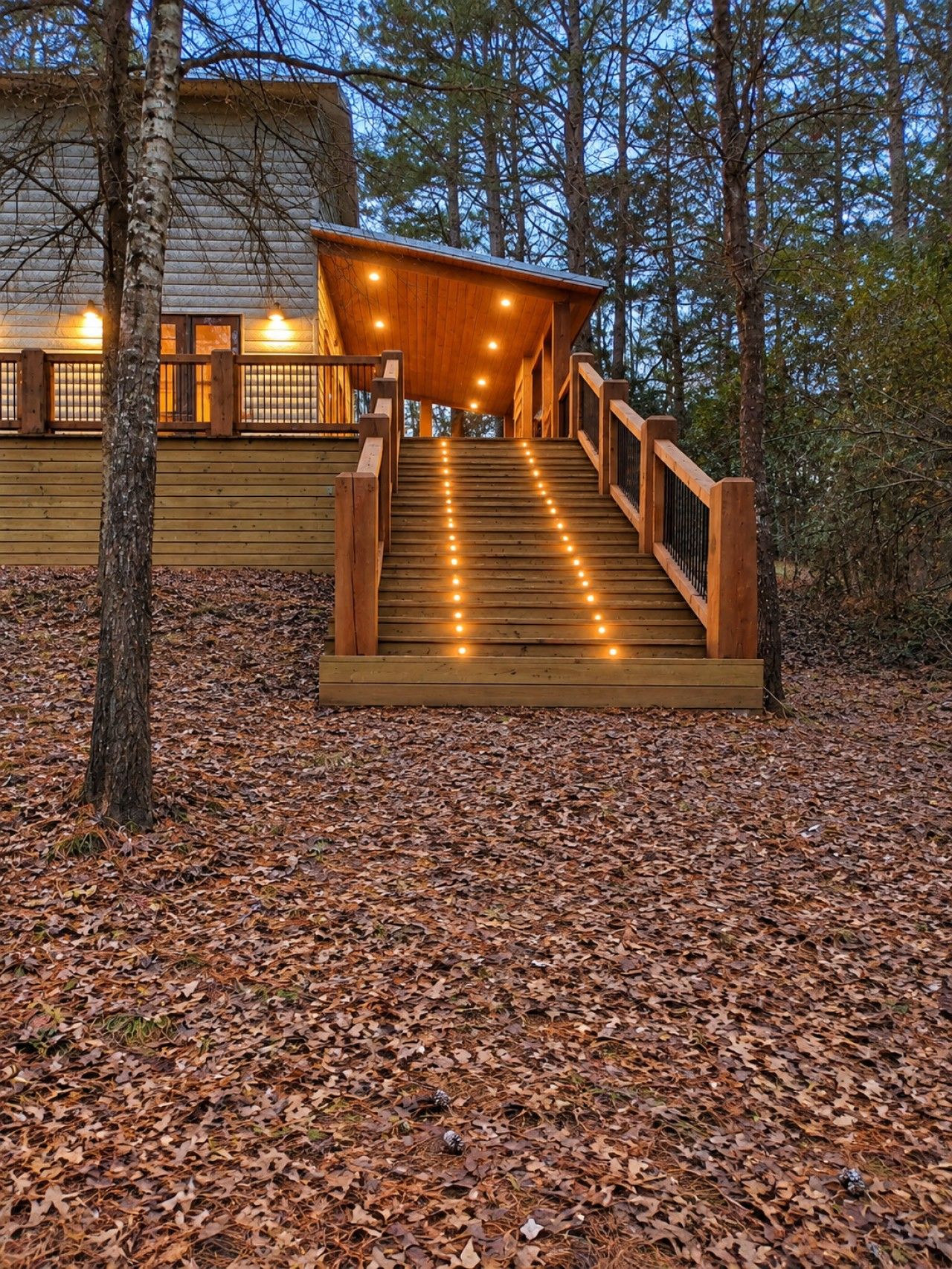A wooden staircase leading to a covered area with lights, surrounded by trees and fallen leaves.