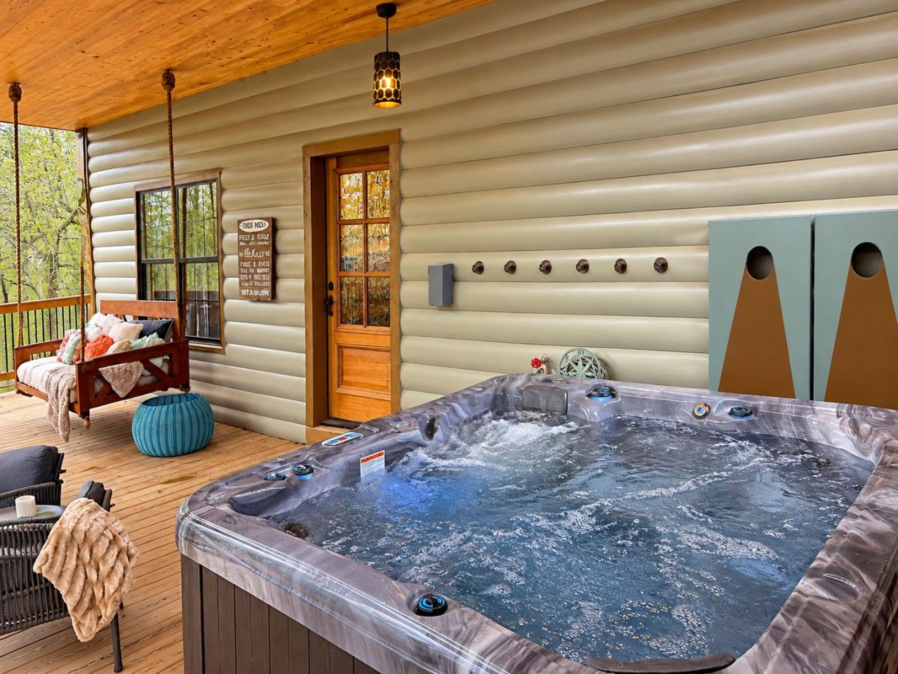 Un jacuzzi en una terraza de madera con una silla y un banco, una puerta y un ventanal con vistas a árboles.
