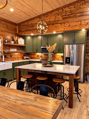 A wooden cabin interior with a central island, bar stools, and a stainless steel refrigerator. - Lofted Pines, Hot Tub, Foosball, Air Hockey, Corn Hole, Horse Shoes, Table Arcade, Tetherball (Broken Bow)