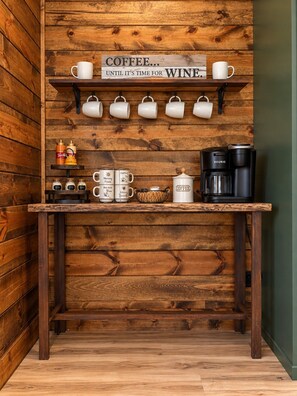 A wooden wall with a sign that reads 'COFFEE... UNTIL IT'S TIME FOR WINE.' Above the sign, there are six white mugs hanging on hooks. Below the sign, there is a coffee maker and a coffee pot on a wooden table. - Lofted Pines, Hot Tub, Foosball, Air Hockey, Corn Hole, Horse Shoes, Table Arcade, Tetherball (Broken Bow)