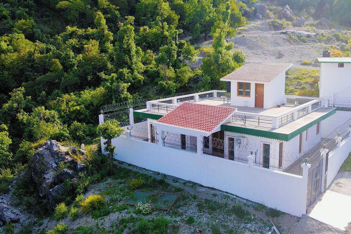 A white building with a red-tiled roof, surrounded by greenery and rocky terrain.
