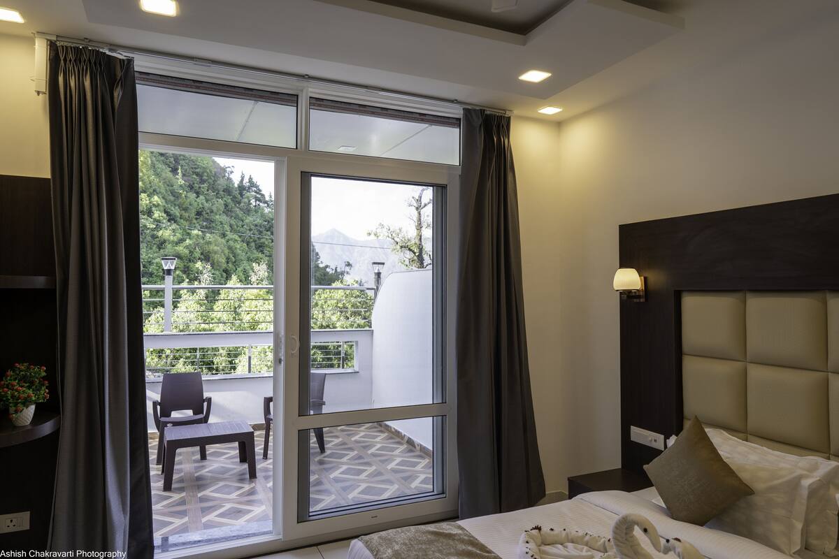 A hotel room with a large sliding glass door leading to an outdoor balcony. The room features a bed with white bedding, a dark headboard, and a nightstand with a lamp.