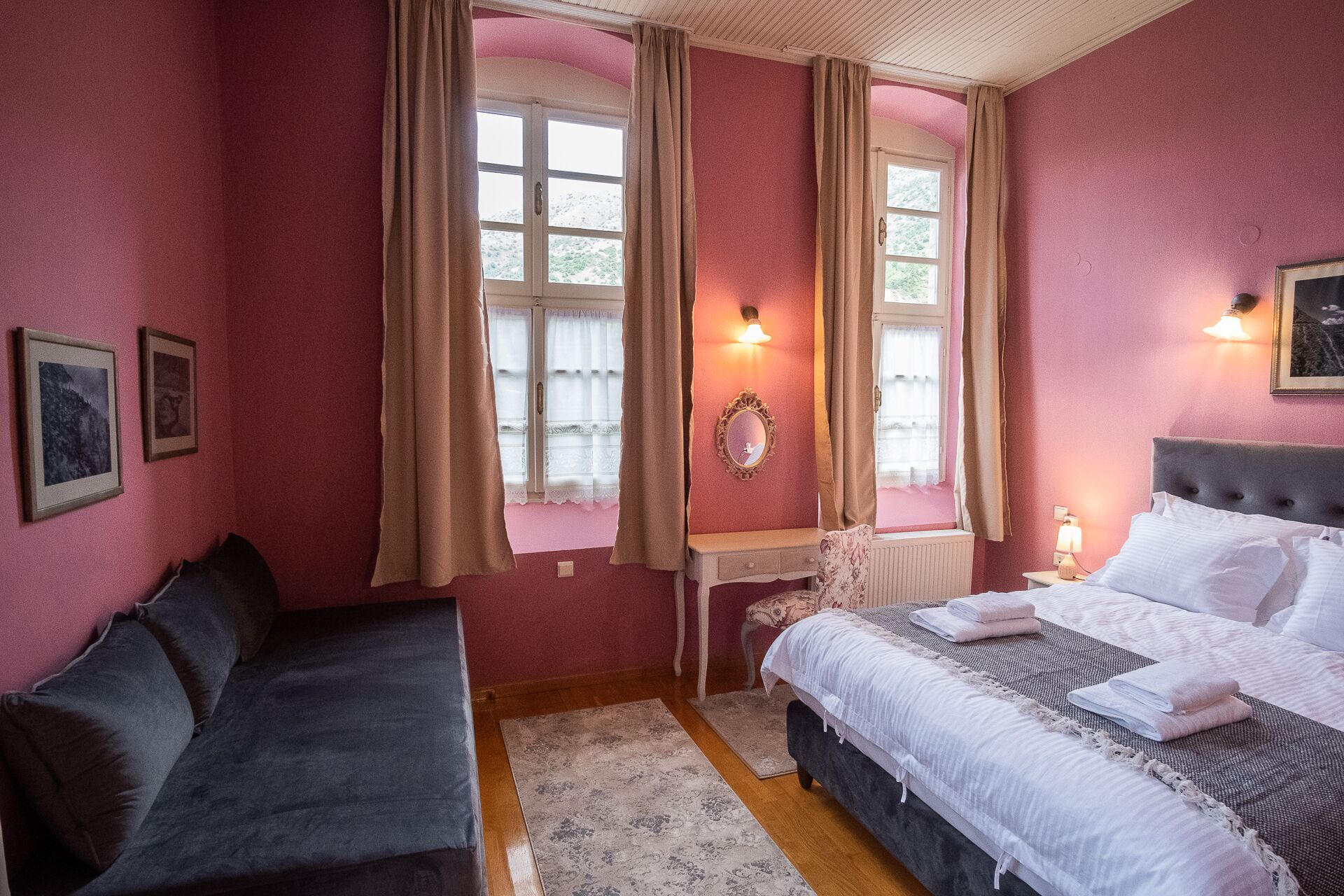 A room with a bed, a sofa, and a desk. There are two windows with curtains.