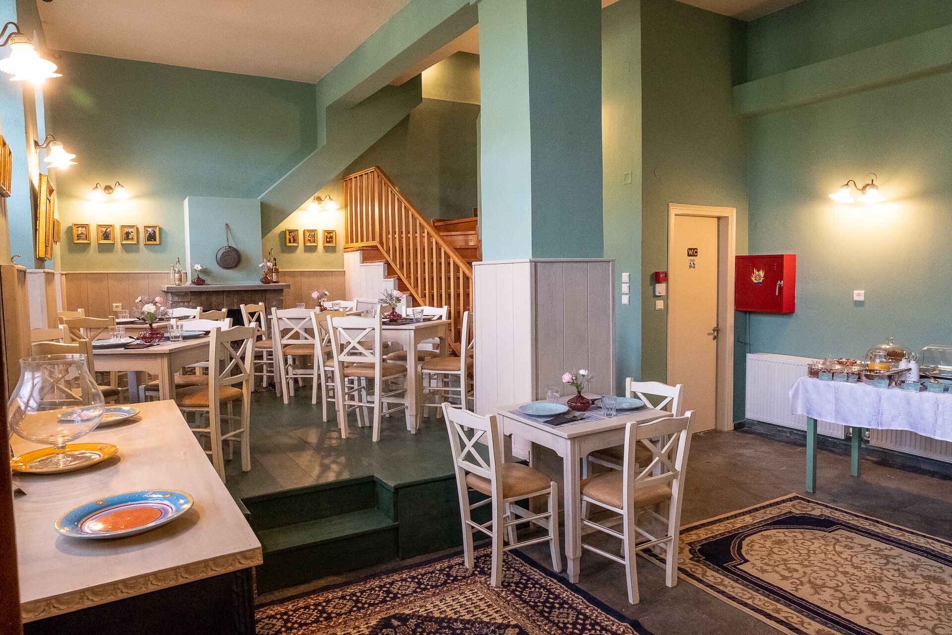 A dining area with multiple tables and chairs, a staircase, and a red emergency box on the wall.