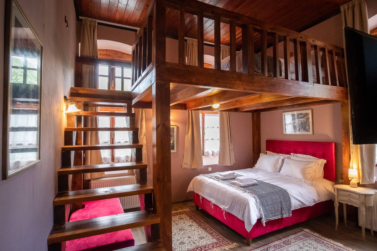 Une chambre avec un lit rouge, une échelle en bois et une fenêtre avec des rideaux.