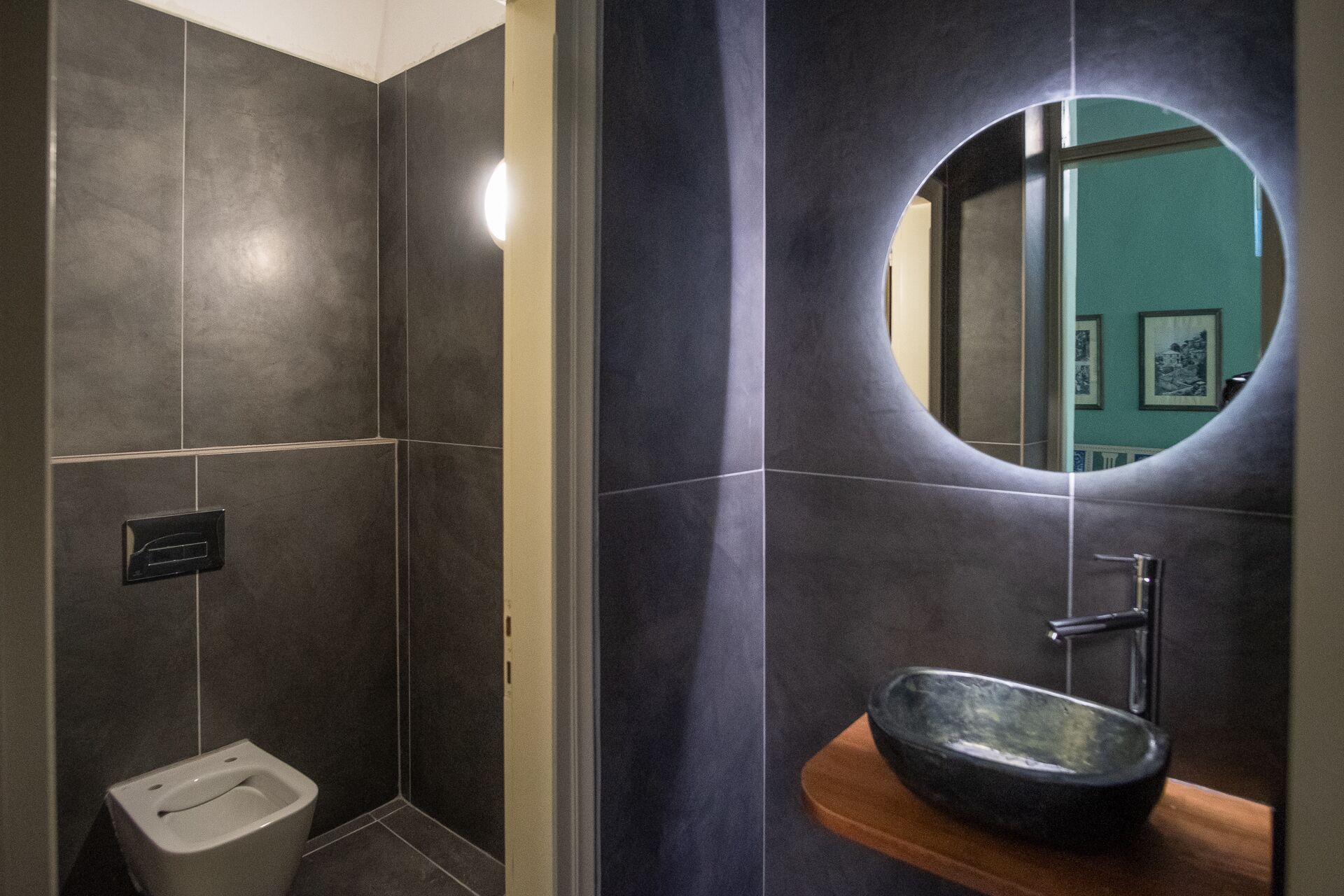 A modern bathroom with dark tiles, a toilet, a sink with a bowl, and a mirror.