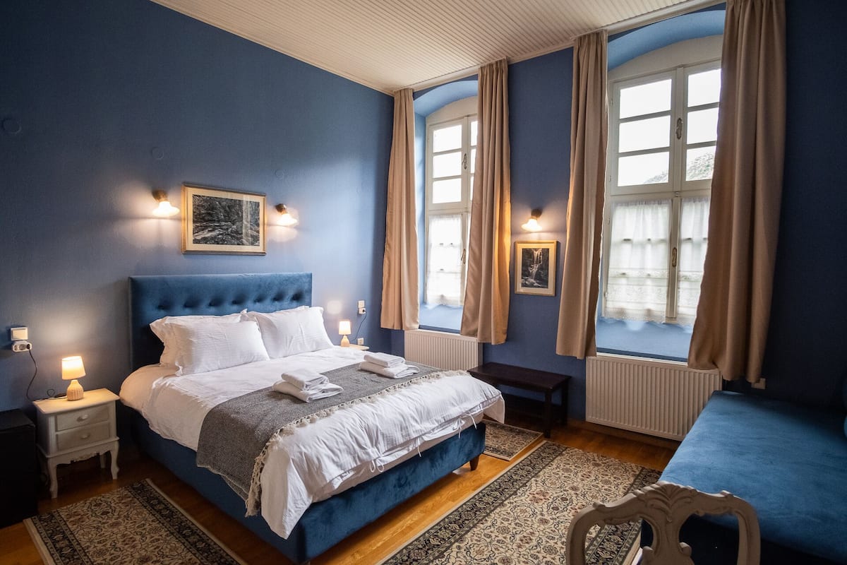 Une chambre à coucher avec un grand lit, deux tables de chevet, une commode, un banc et une fenêtre avec des rideaux.