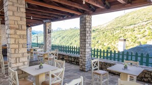 Eine Terrasse im Freien mit steinernen Säulen, Holztischen und -stühlen mit Blick auf einen grünen Hügel.