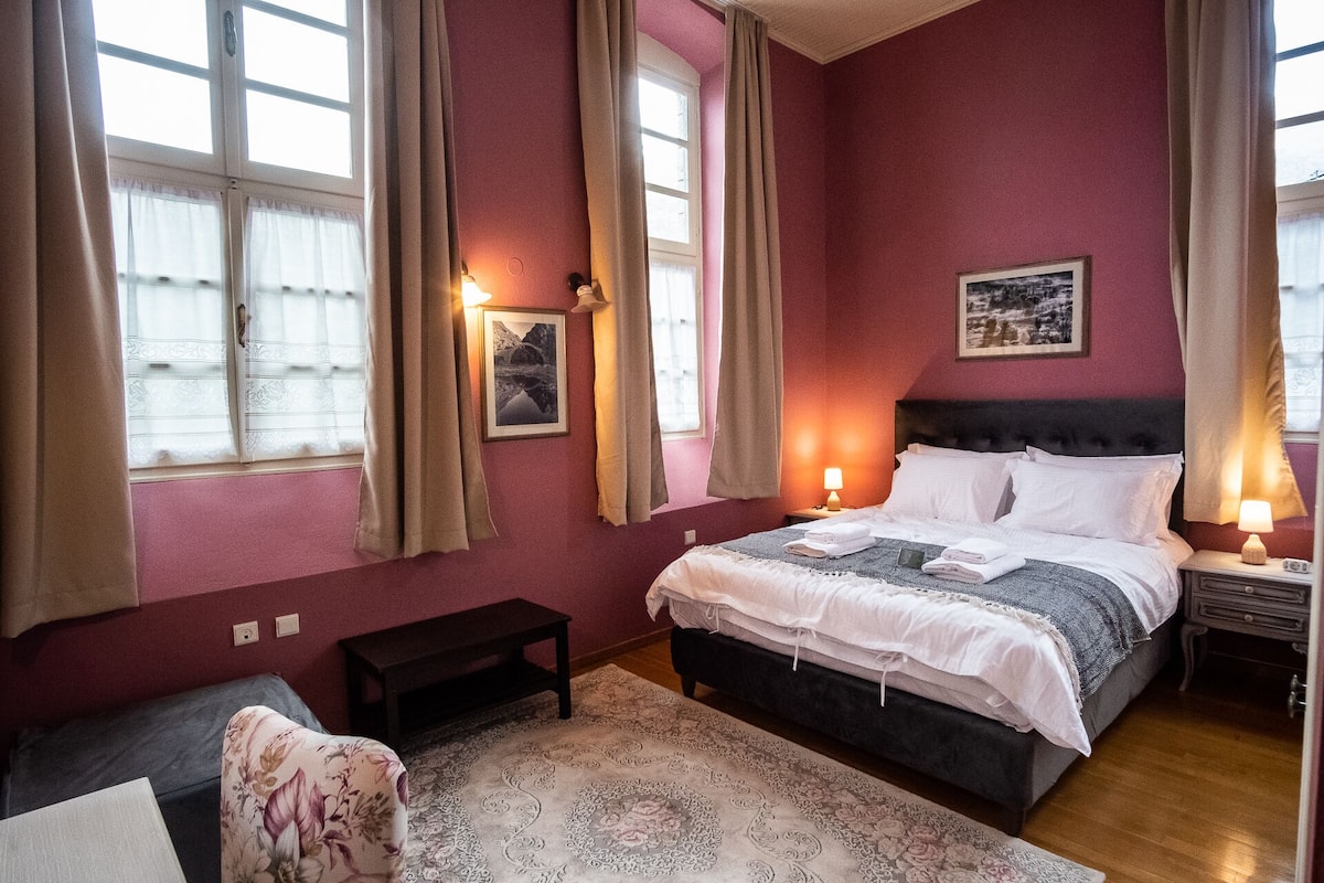 Une chambre à coucher comprenant un lit, des tables de chevet, une commode, une petite table, un banc et une fenêtre avec des rideaux.
