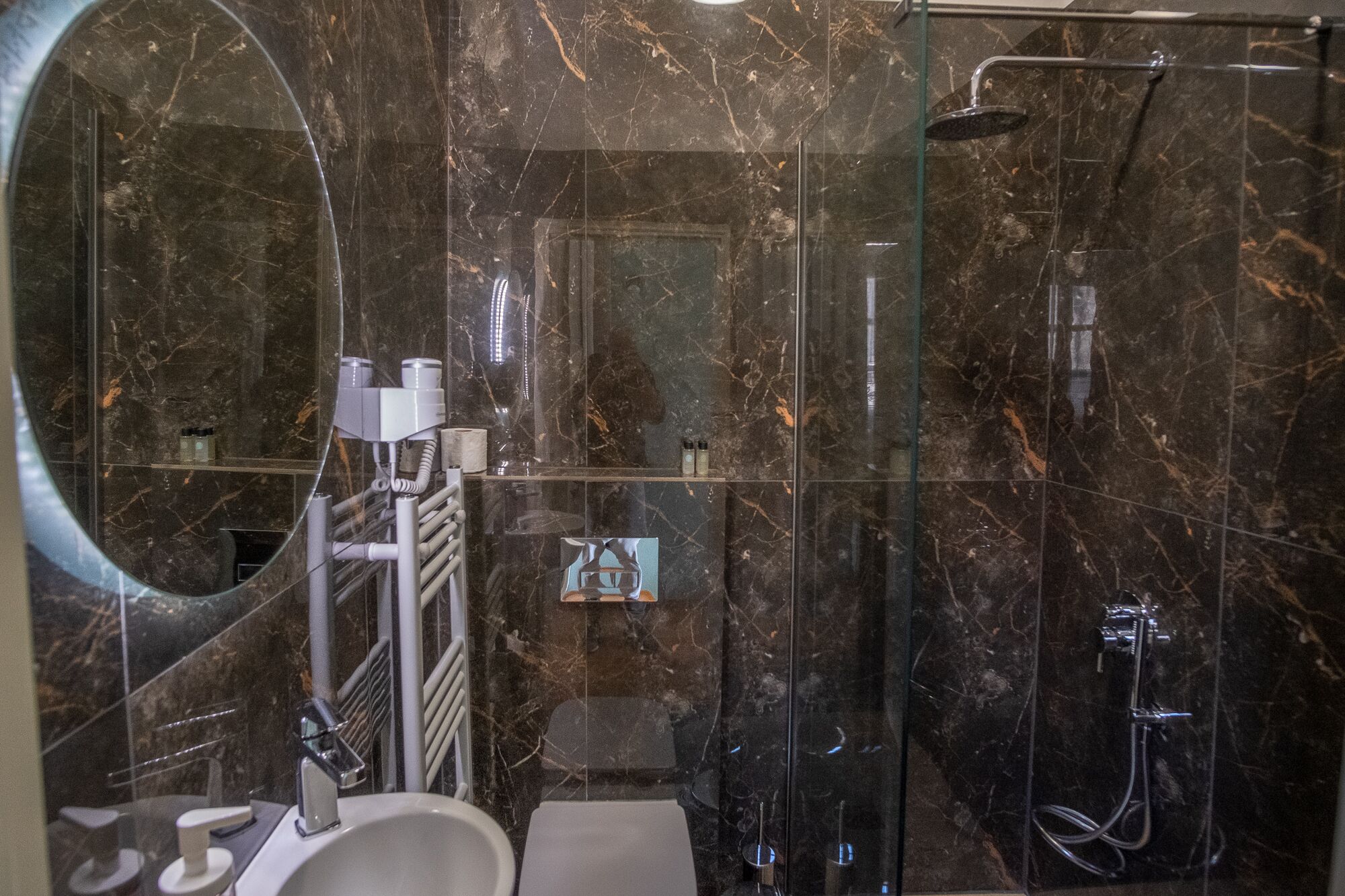 A modern bathroom with a large mirror, a sink, a glass shower enclosure, and marble walls.