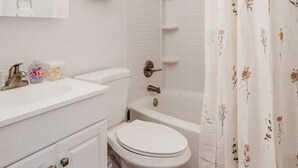 A bathroom with a white toilet, a sink with a mirror, and a shower with a floral curtain. - Cozy, Central 1B Apt Close to UC Hospitals, DT (Norwood)