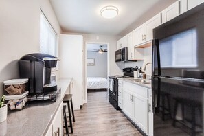 A compact kitchen with a refrigerator, microwave, and stove. There is a small dining area with stools and a coffee machine. A bed is visible in the background.