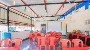 Un salón de conferencias con sillas y mesas de plástico rojo, una pantalla de proyector y una zona de buffet con recipientes de comida.