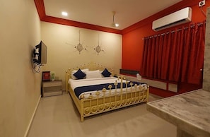 A bedroom with a bed, bedside table, nightstand, wall-mounted TV, air conditioning unit, and a red curtain. - Hotel Suraj inn (Surat)