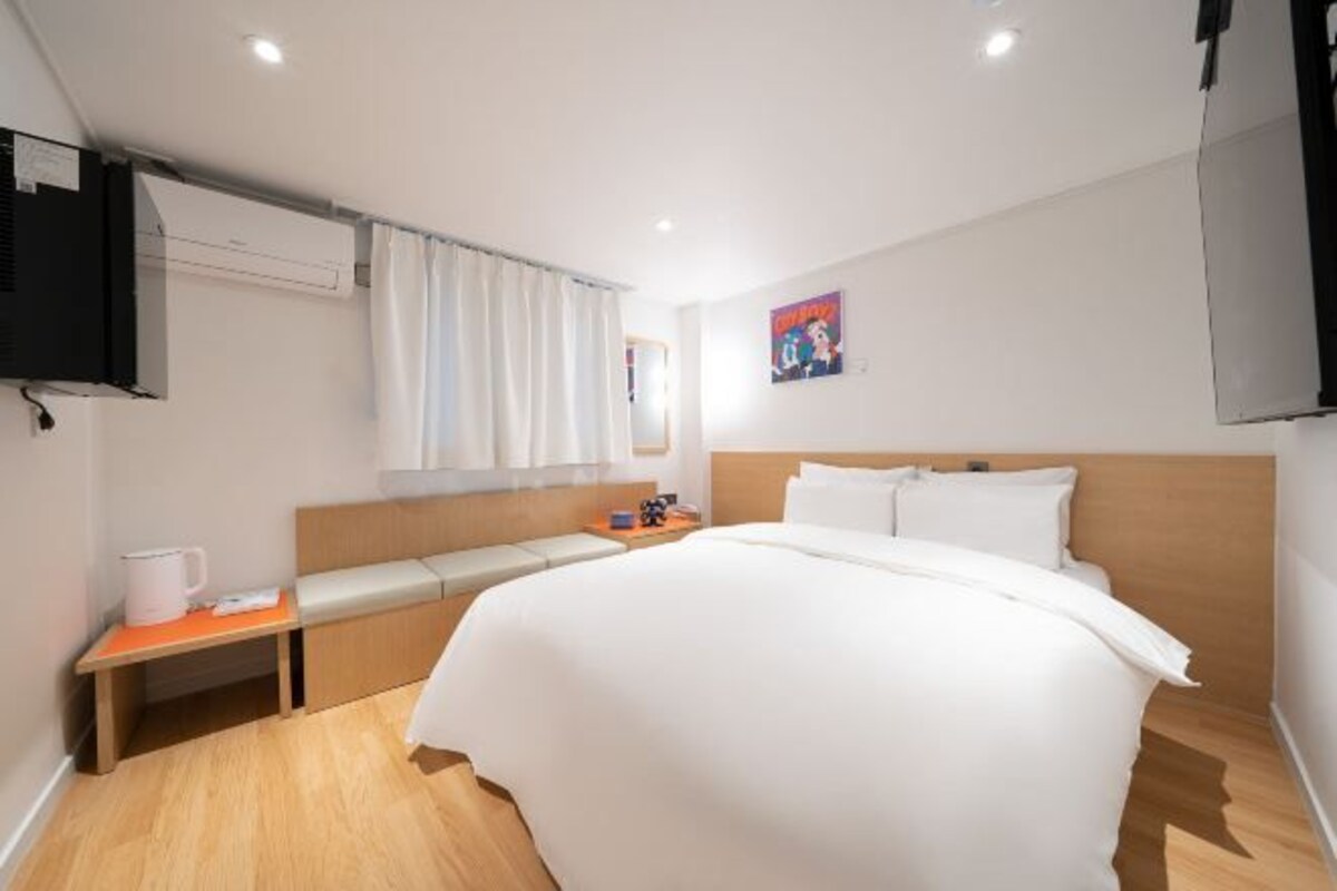 Une chambre d’hôtel avec un grand lit, une table de chevet, un téléviseur fixé au mur et une fenêtre avec des rideaux.