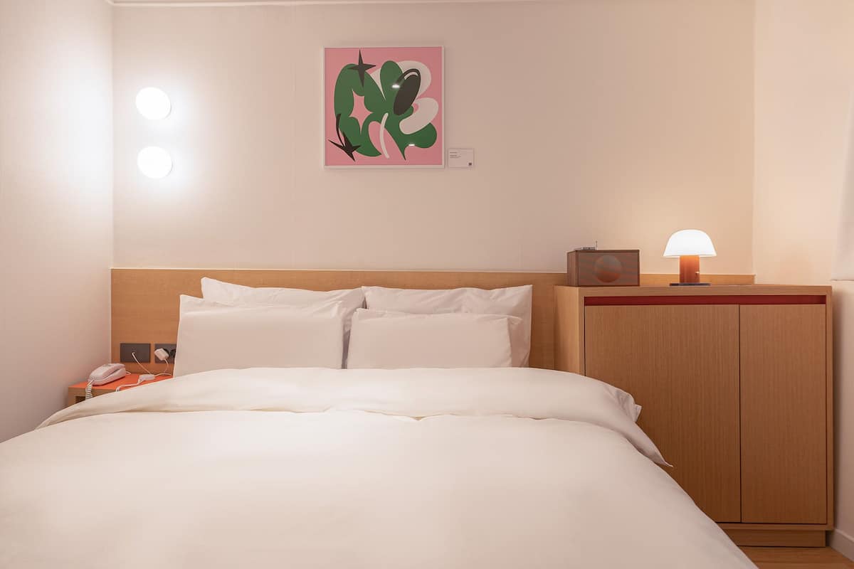Ein Hotelzimmer mit Bett, Nachttisch, Lampe und einem Kunstwerk an der Wand.