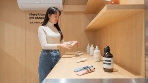 Eine Frau in Beige-Pullover und Jeans interagiert mit Hautpflegeprodukten auf einer Holzauskładung in einem gut beleuchteten Raum, in dem ein Klimagerät steht und ein Schild an der Wand hängt.