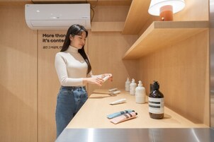 Una mujer con suéter color beige y jeans interactúa con productos para el cuidado de la piel sobre una encimera de madera en una habitación bien iluminada, con un aire acondicionado y un cartel en la pared.