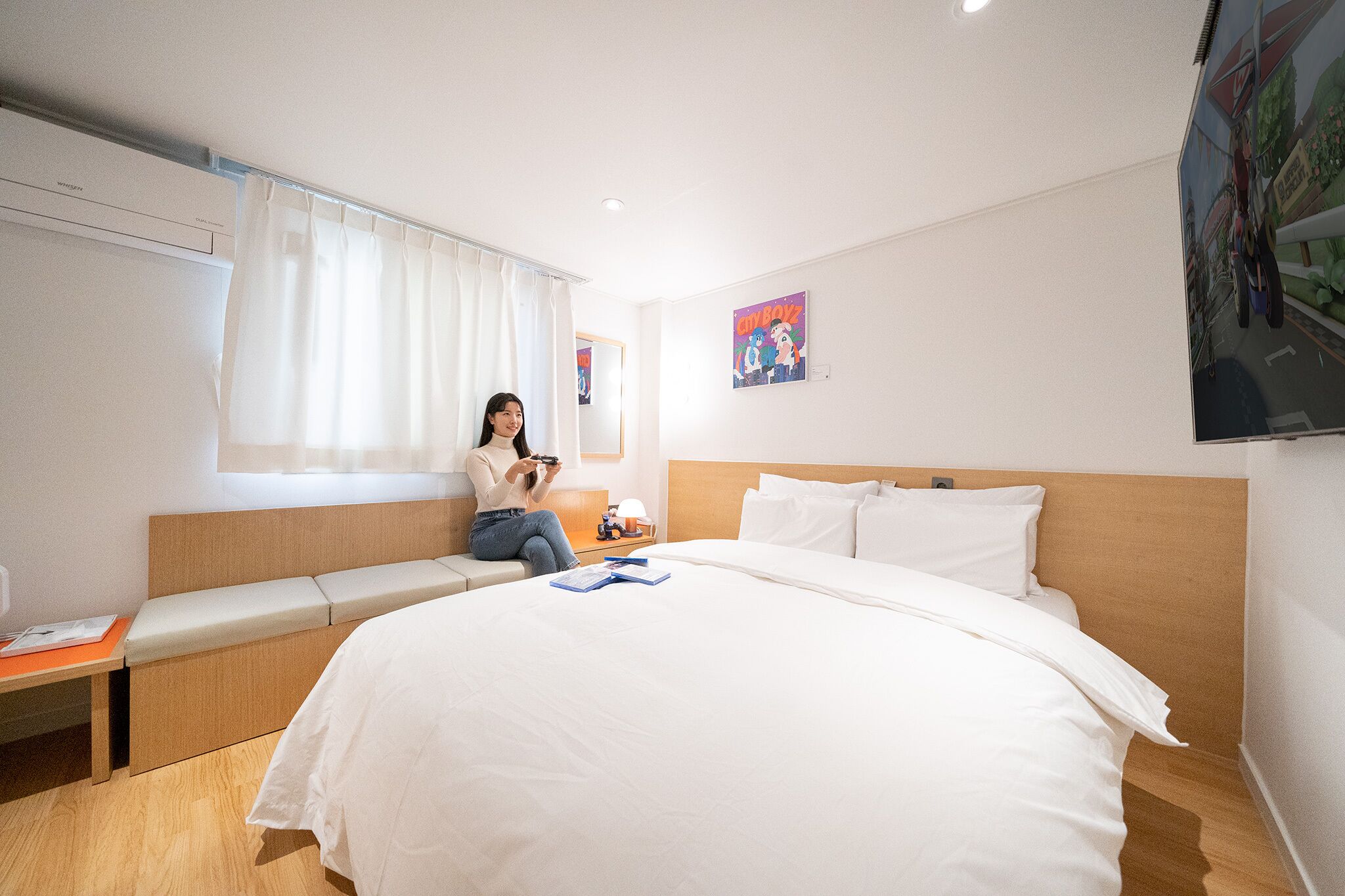 A hotel room with a large bed, a bedside table, a window with curtains, a wall-mounted air conditioner, and a poster on the wall.