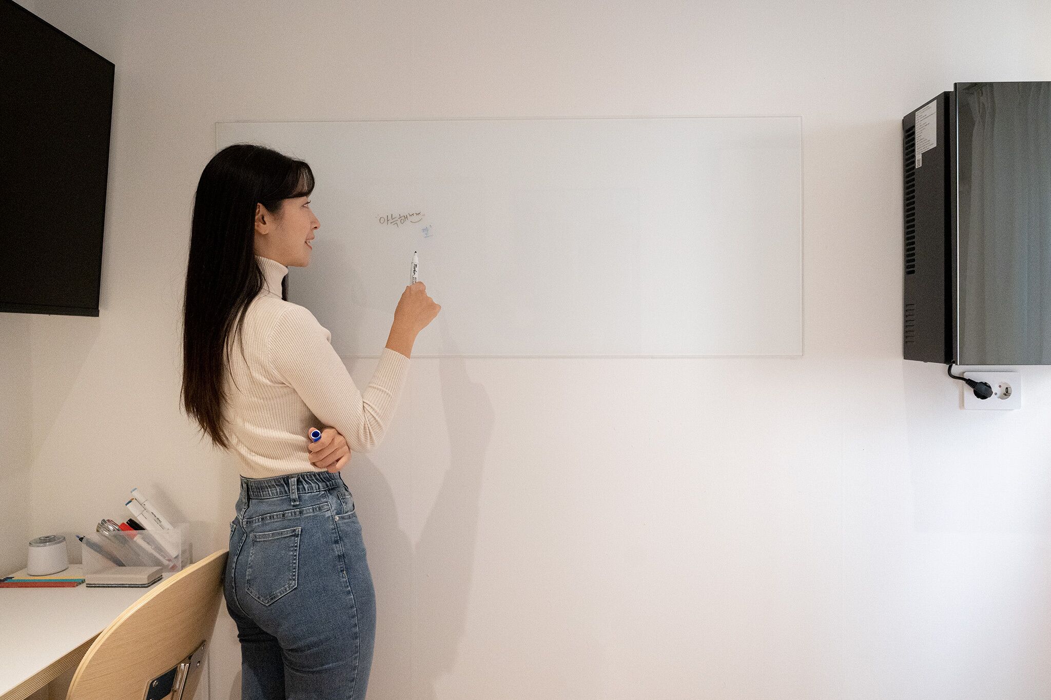 A person standing in front of a whiteboard in a room with a desk and a black cabinet.