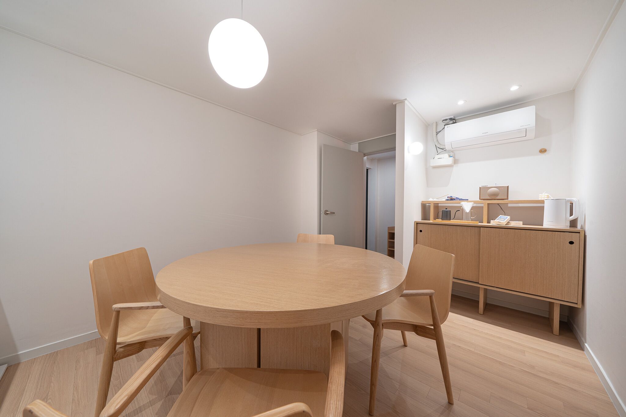 Ein moderner, gut beleuchteter Raum mit einem runden Holztisch, umgeben von Stühlen, einer Holzkommode mit Spiegel und einer Deckenleuchte.