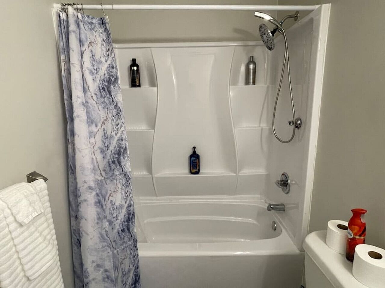A bathroom with a bathtub, shower, and toilet. There are toiletries on the shelves and a towel rack.