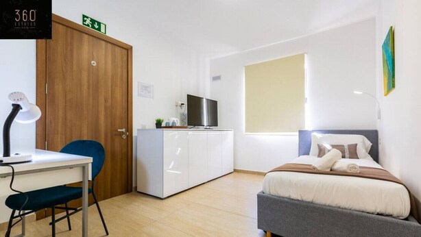 A modern bedroom with a bed, a desk with a chair, a wardrobe, a television, and a window. - Stylish 1BR Meridian in central St Julians (San Ġiljan)