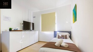 A modern hotel room with a bed, a TV on a white cabinet, a window with blinds, and a painting on the wall. - Stylish 1BR Meridian in central St Julians (San Ġiljan)