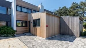 A modern building with a combination of wooden and grey exterior panels, a small wooden structure, and a paved entrance area. - Holiday Home by the Sea and Dunes (Nieuwvliet-Bad)