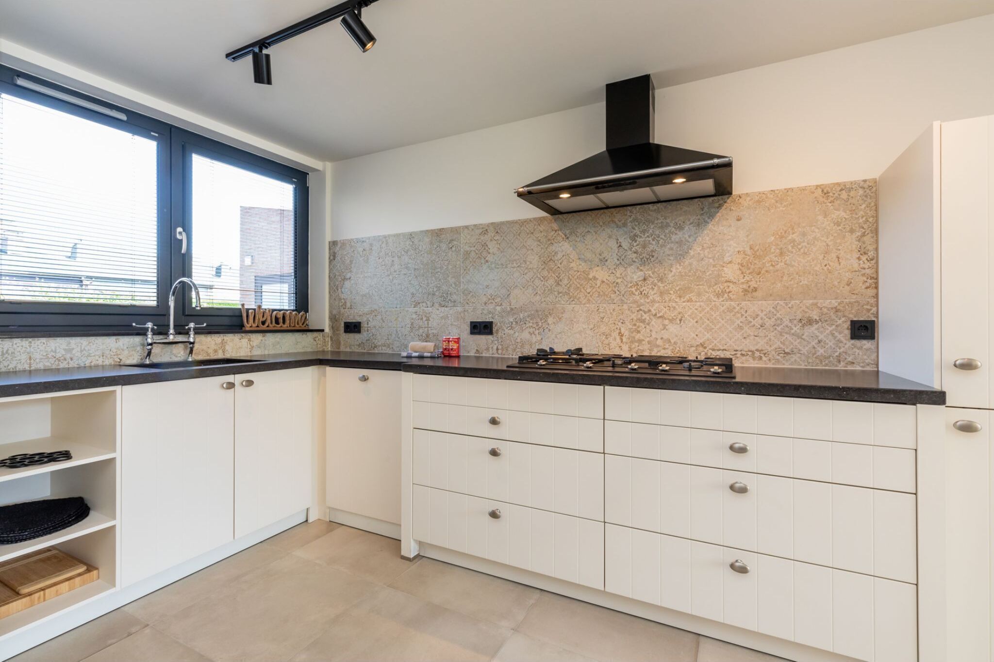 Een moderne keuken met witte kasten, een zwart aanrecht, een gasfornuis en een raam met jaloezieën.
