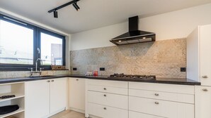 A modern kitchen with white cabinets, a black countertop, a gas stove, and a window with blinds. - Holiday Home by the Sea and Dunes (Nieuwvliet-Bad)
