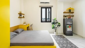 Ein Schlafzimmer mit einem Bett, einem Schreibtisch, einem Stuhl, einem Fenster, einer Klimaanlage, einem Kühlschrank und einer Zimmerpflanze.
