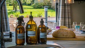 A kitchen counter with bottles of olive oil, a loaf of bread, and a sink with a garden view.