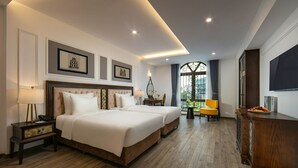 A hotel room with two beds, a wooden floor, a desk with a lamp, a chair, and a view of greenery outside. - Gloud Hotel (Hanoi)