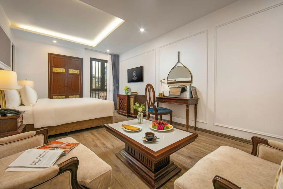 A hotel room with a bed, a desk with a chair, a coffee table with a plate of fruit, and a lamp.