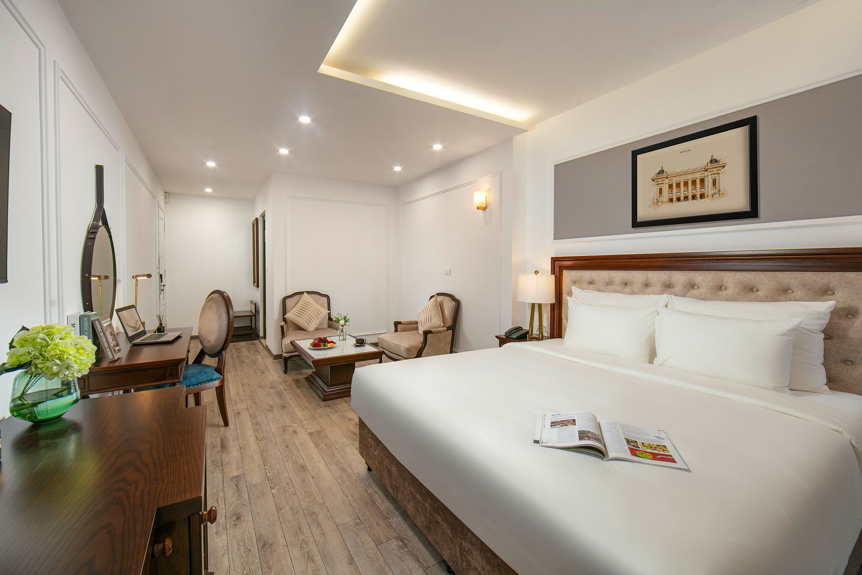 A hotel room with a large bed, a desk with a laptop, two chairs, a small table with a vase, and a framed picture on the wall.