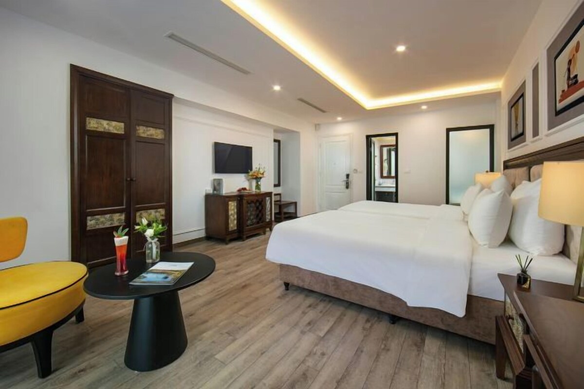A hotel room with a large bed, a small round table, a yellow armchair, a wooden wardrobe, and a television on the wall.