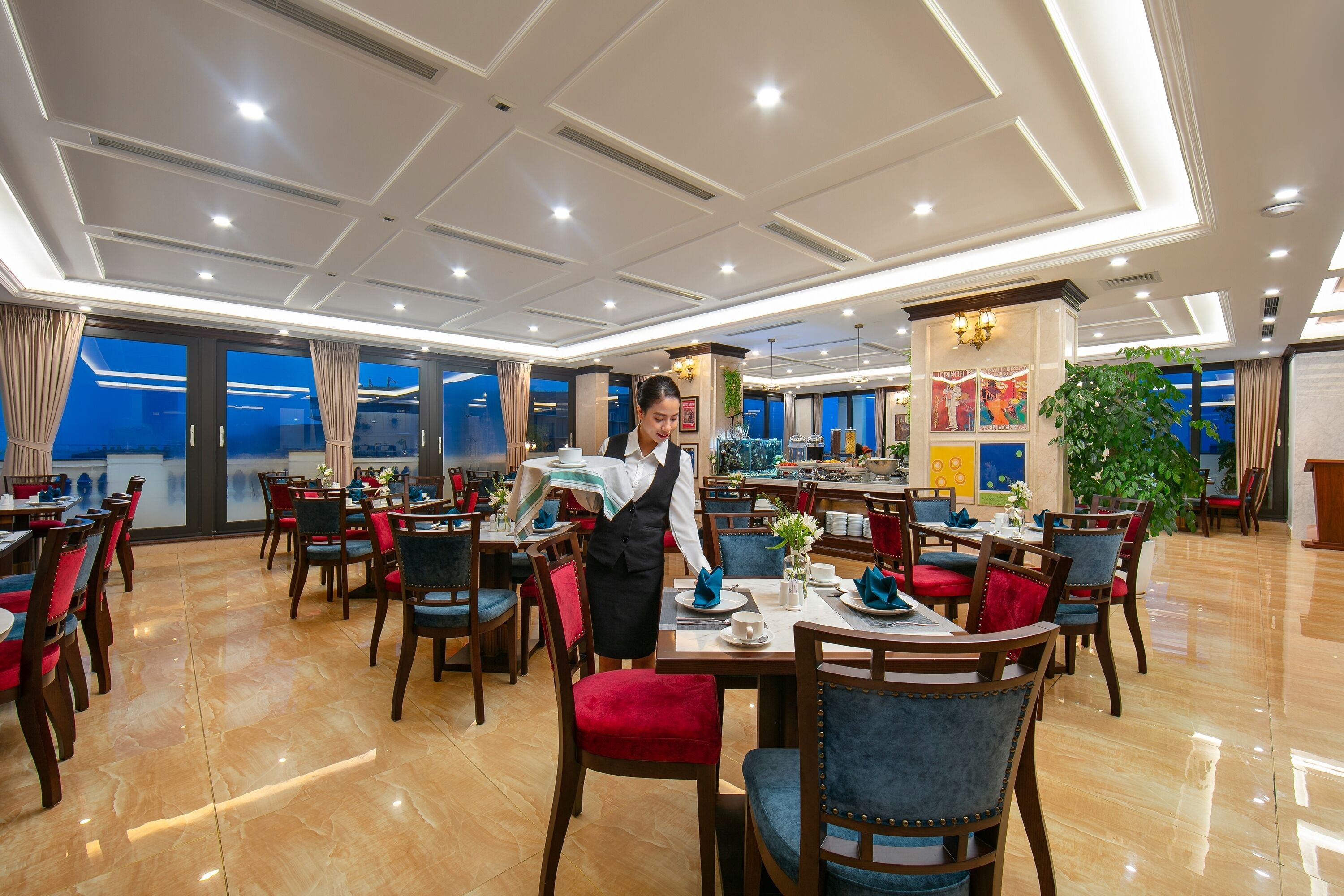 A spacious hotel dining area with multiple tables and chairs, a well-lit ceiling, and large windows offering a view.