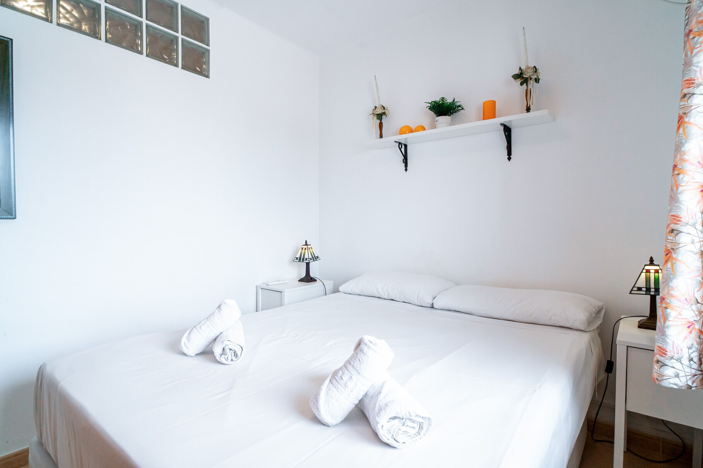 A neatly made bed with white linens and rolled towels, a bedside table with a lamp, a wall-mounted shelf with decorative items, and a window with blinds.