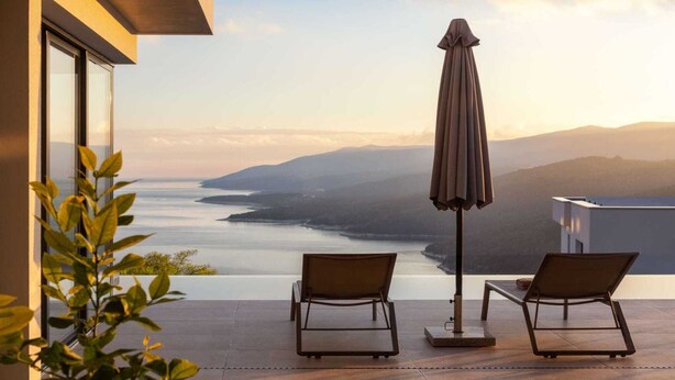 A balcony with two chairs and a table, overlooking a body of water and distant mountains.