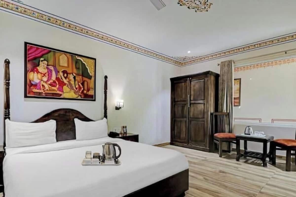 Habitación de hotel con cama, escritorio, dos sillas, armario y dos cuadros en las paredes.