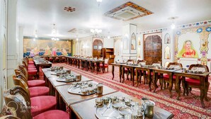 Une salle à manger traditionnelle avec de longues tables dressées pour le repas, agrémentée de décorations murales complexes et d’un tapis à motifs.