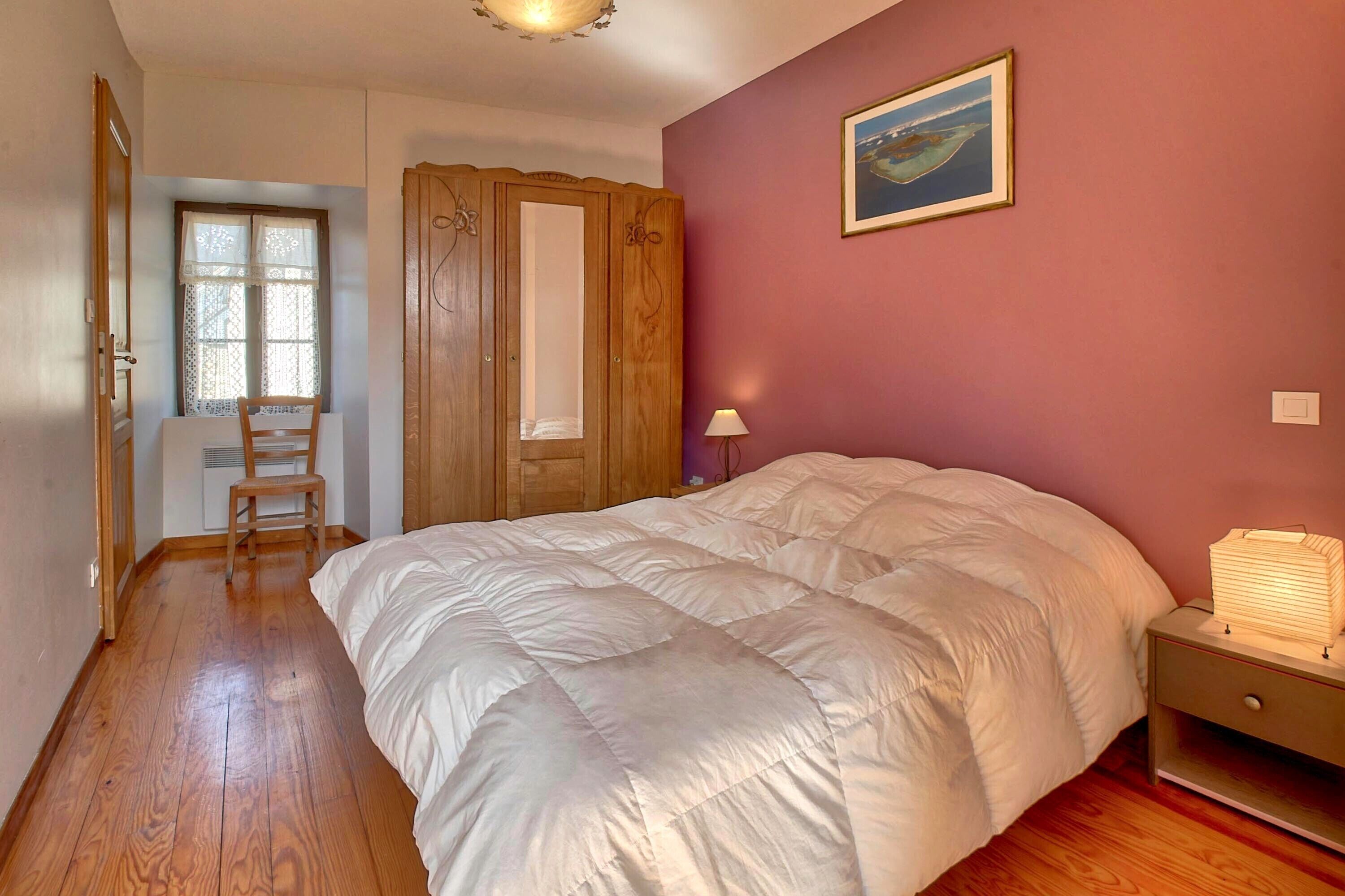 A bedroom with a large bed, a wooden wardrobe, a chair, a bedside table with a lamp, and a painting on the wall.