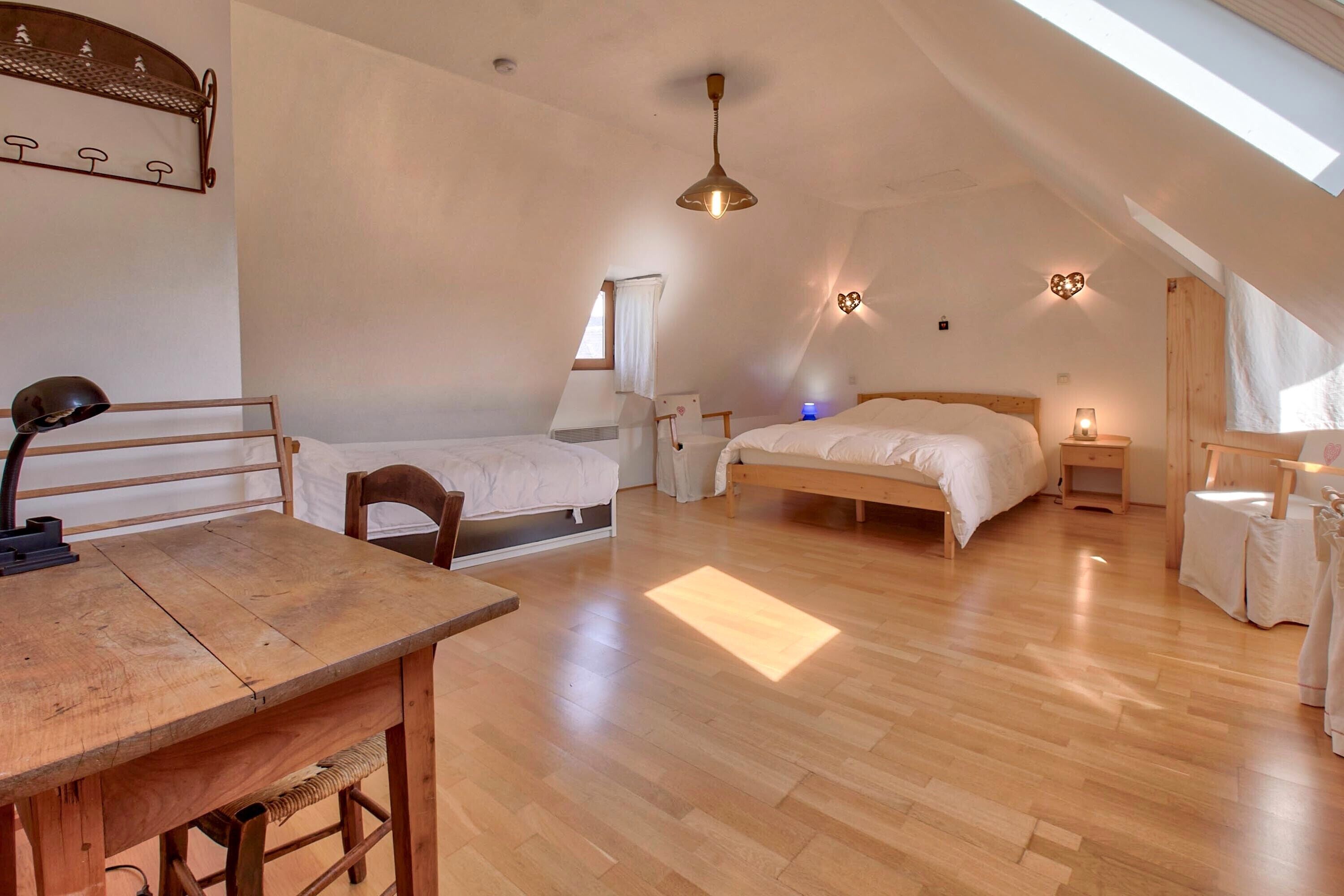 A bedroom with a sloped ceiling, a wooden table, two beds, a hanging light fixture, and a wall-mounted shelf.