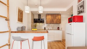 Une cuisine moderne avec un îlot central en bois, deux tabourets de bar, un réfrigérateur blanc et un téléviseur fixé au-dessus de l’îlot.