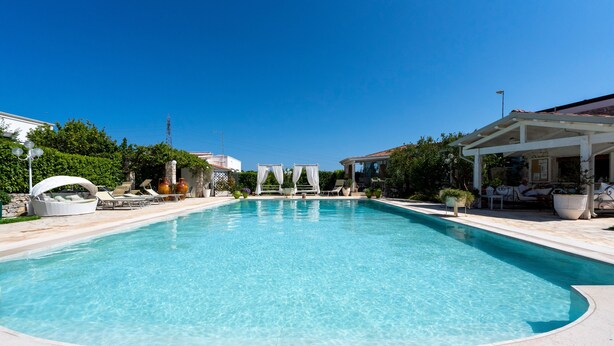 A clear swimming pool with lounge chairs and umbrellas, surrounded by greenery and buildings.