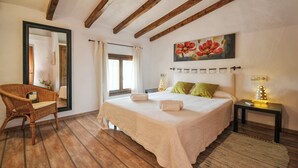 A bedroom with a large bed, a wicker chair, a painting, and a nightstand with a lamp. - Country House 'Cadernera' with Private Pool, Wi-Fi and Air Conditioning (Búger)